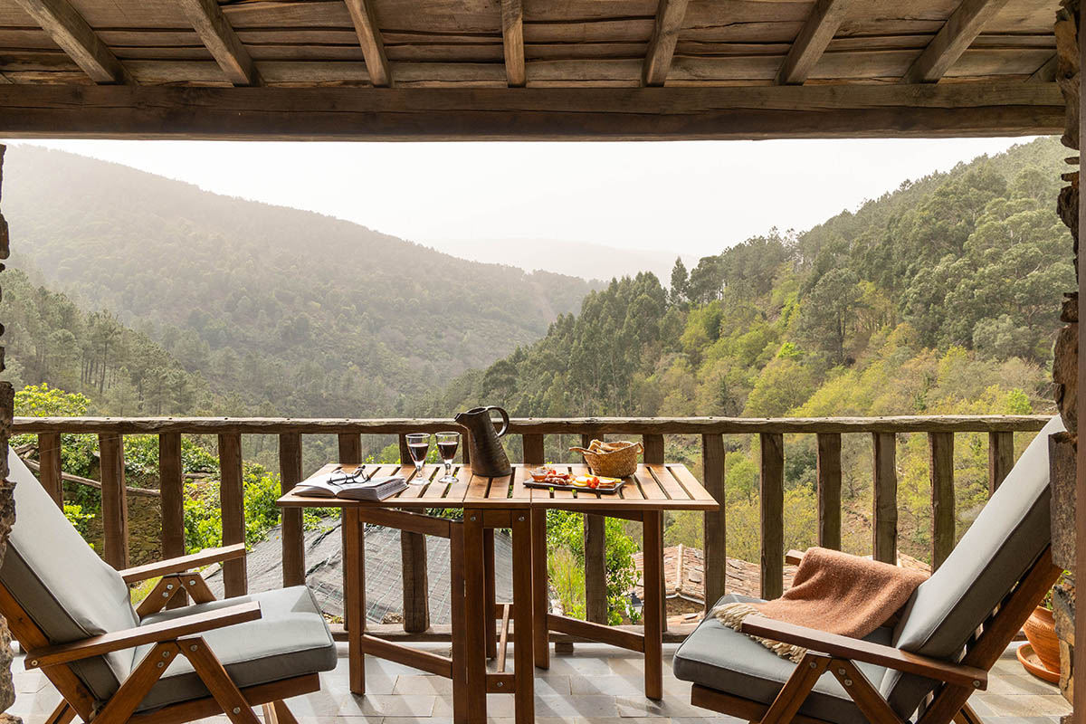 A wooden balcony at the Cerdeira retreat: two chairs and a small table set with wine glasses, looking out across forested mountain valleys in the Serra da Lousã at golden hour.