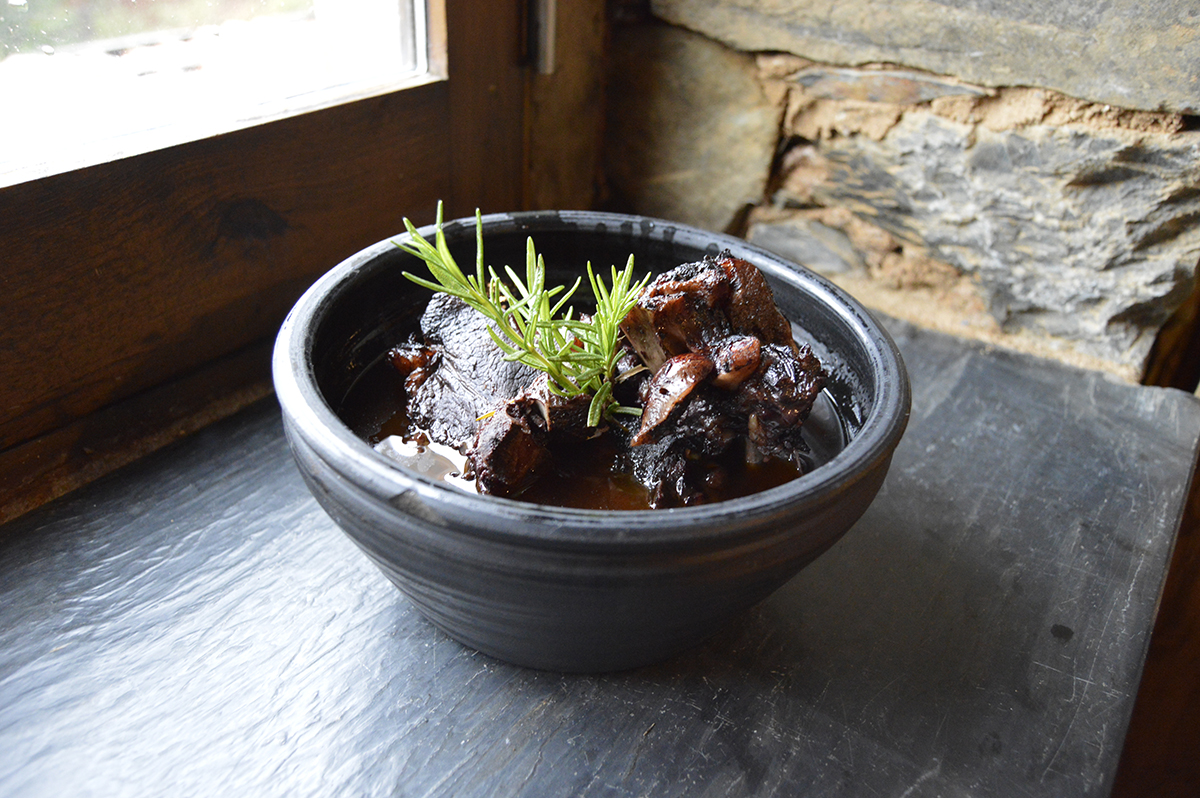 A bowl of slow-braised meat with fresh rosemary on a slate counter, lit by morning window light against a stone wall — a Cerdeira chef's table.
