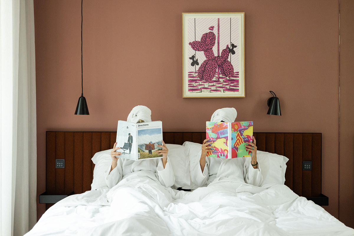A guest bedroom at the Lisbon boutique hotel: terracotta walls, two figures in white robes reading in bed, framed contemporary art above the headboard.