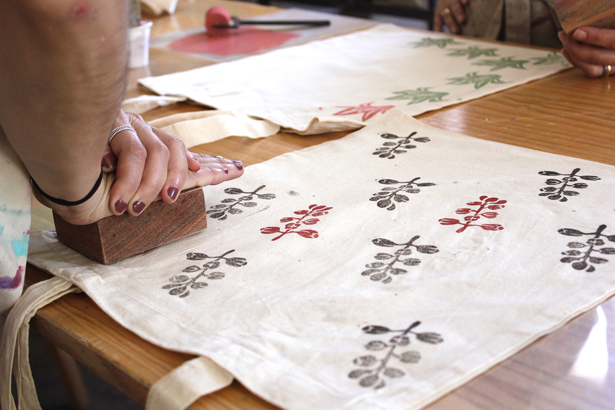 Hands pressing a hand-carved wooden block to print rows of botanical leaves in red, green, and black pigment onto a cream linen panel.
