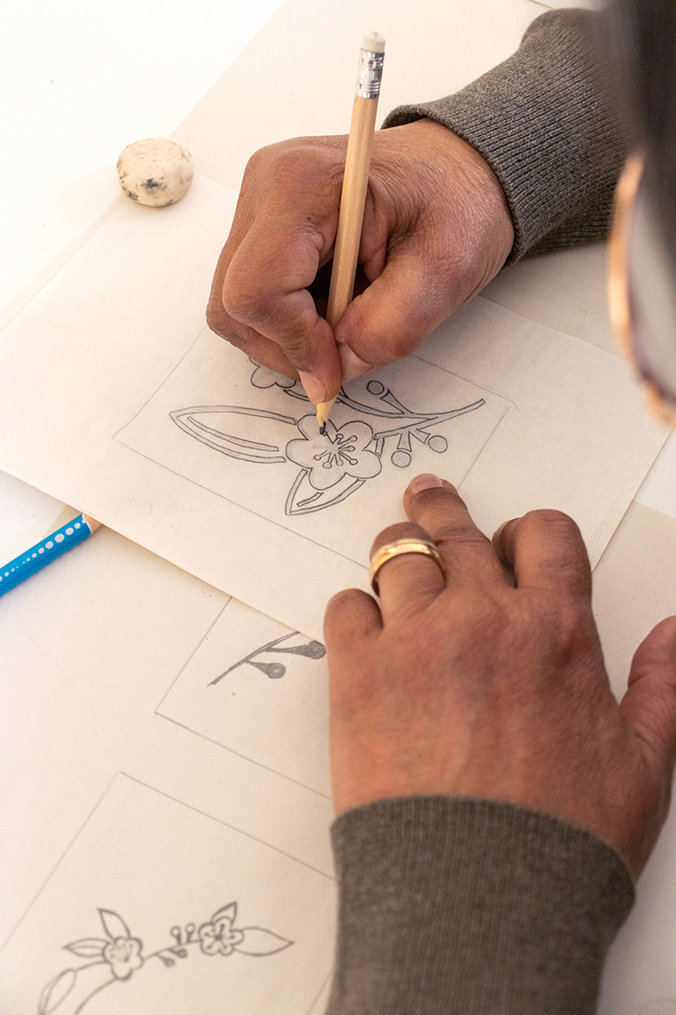 A workshop participant sketching botanical and floral designs in pencil on paper, preparing pattern studies for a hand-carved block.